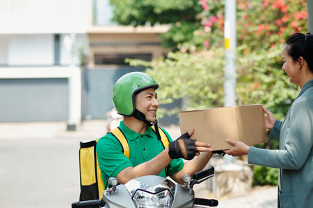 Smiling courier sitting on scooter and giving box to customerの写真素材