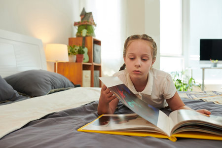 Girl resting on bed and turning pages of book of photosの写真素材