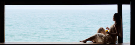 Barefoot young woman in silk flowing dress relaxing by coast with glass of fruit cocktailの写真素材