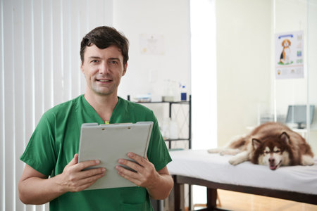 Portrait of happy veterinarian with clipboard standing in office cleanic, samoyed dog in background