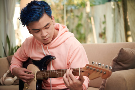 Concentrated creative young man with blue hair playing guitar ta home
