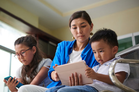 Mother and kids watching entertaining videos on devices when waiting in queue in hospitalの写真素材