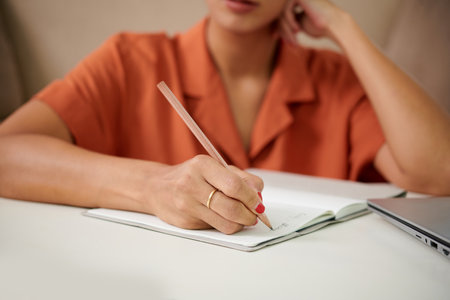Close-up of young woman making notes in copybook while sitting at table in the roomの写真素材