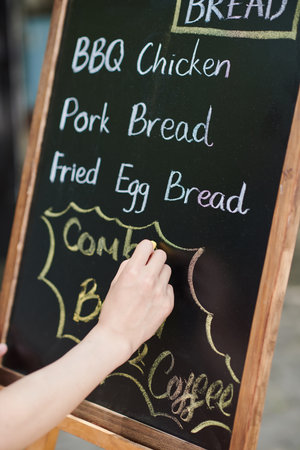 Hand of young female vendor of street fast food writing down menu on blackboard with white chalk at the beginning of working dayの写真素材
