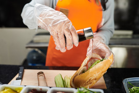 Gloved hands of young female worker of street fast food truck pouring sauce into appetizing hotdog with variety of ingredients insideの写真素材