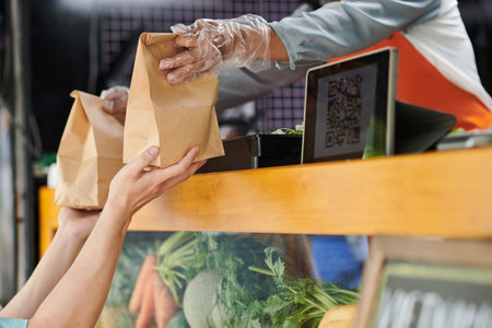Hands in protective gloves of young female vendor passing paper package with sandwiches or hotdogs...