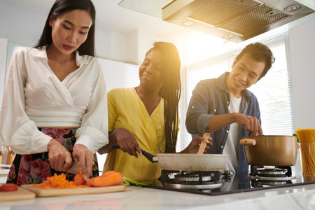 Young black woman standing by gas stove in the kitchen, mixing ingredients in frying pan and looking at her friend chopping fresh carrotの写真素材