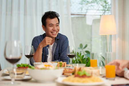 Young cheerful Asian man in casualwear sitting by served table at dinner prepared for guests and...