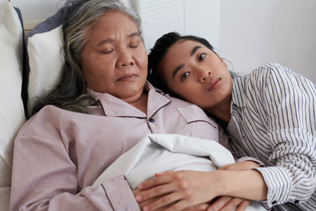 Pensive serious young woman leaning on shoulder of sick sleeping motherの写真素材