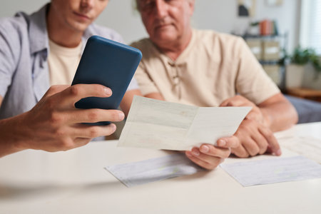 Young man helping grandfather to pay utility bills via mobile applicationの写真素材