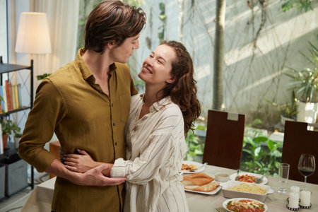 Young couple in love standing at dinner table at home, hugging at looking at each otherの写真素材