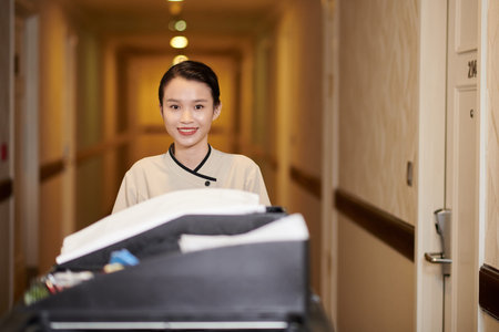 Young hotel maid pushing trolley with dirty linen and cleaning detergents