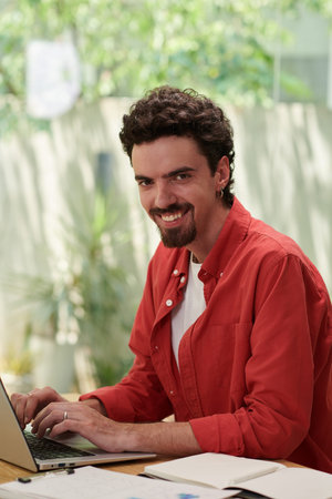 Smiling software developer in bright red shirt working on laptop in home office