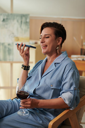 Cheerful mature woman drinking wine and recording voice message for friendの写真素材