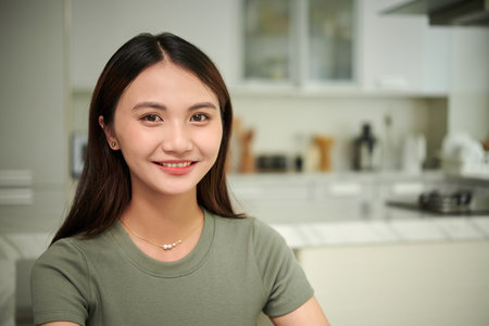 Portrait of cheerful young Asian woman in t-shirt standing in kitchenの写真素材