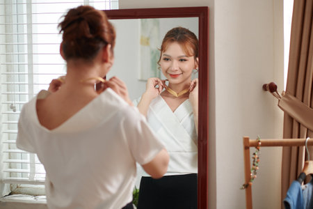 Smiling young transgender woman putting on jewelry in front of mirrorの写真素材