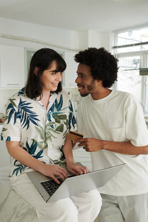 Happy young man with credit card looking at his wife or girlfriend using laptop while consulting...