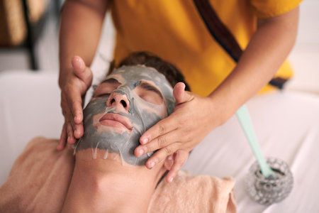 Focus on hands of young female beautician spreading hydrating sheet mask on face of relaxed male client with closed eyes visiting spa salonの写真素材