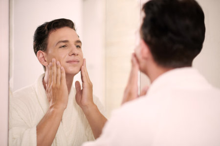 Young multiethnic man in white bathrobe touching his face while standing in front of mirror in bathroom and looking at his reflectionの写真素材