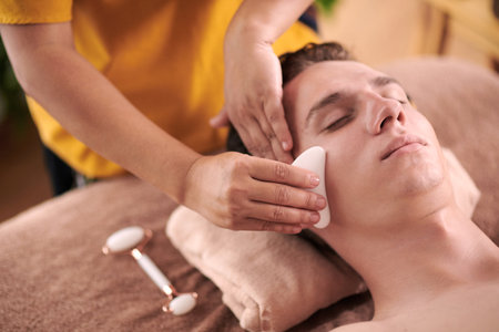 Young female beautician with guasha smoothing skin of male client face during luxurious spa procedure while standing behind his headの写真素材