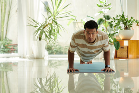 Young active man in striped t-shirt looking at camera while doing push ups on the floor of living room during workout in the morningの写真素材