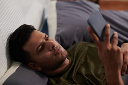 Young man in t-shirt looking through online news, watching video or chatting in smartphone while...