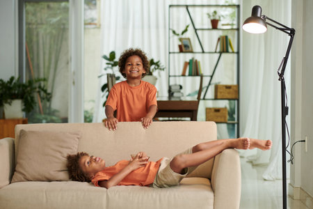 Cheerful kids spending time at home, eating snacks and resting on couchの写真素材