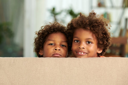 Children with curly hair hiding behind couch in living roomの写真素材