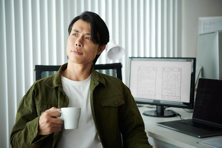 Portrait of pensive inspired software developer with cup of coffee sitting at his desk