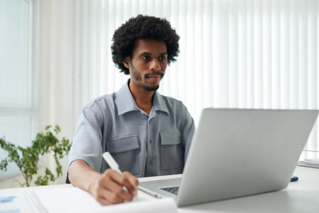 Smiling businessman reading report on laptop screen and taking notes on paper sheetの写真素材