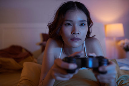 Portrait of young Asian woman lying on bed and playing videogame late at night