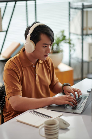 Young Vietnamese tester listening to music when working at desk in home office