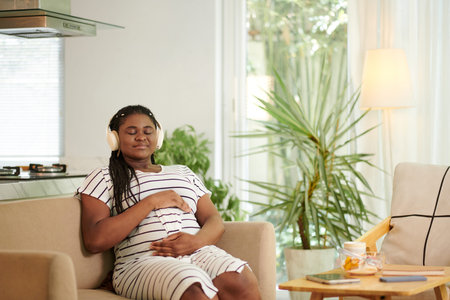 Pregnant Black woman listening to calm music in headphones and touching her bellyの写真素材
