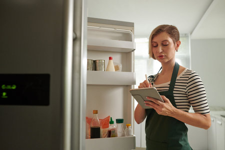 Woman standing in front of opened fridge and filling shopping list on tablet computer