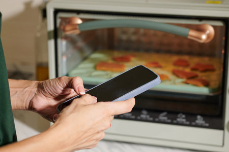 Hands of woman setting time on smartphone after putting tray with cookies in ovenの写真素材