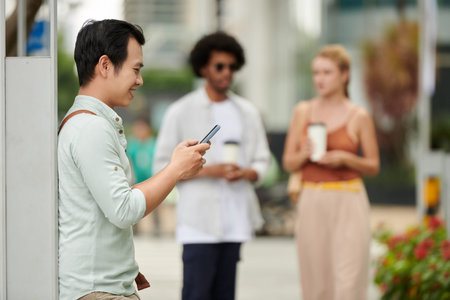 Smiling young Vietnamese man answering text messages before meeting with coworkers