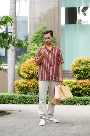 Smiling young man with shopping bags reading delivery notification on smartphone