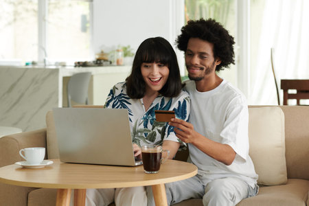 Happy young intercultural couple choosing online goods in front of laptop while African American man...