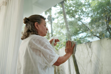 Senior woman cleaning windows in her house with spray detergentの写真素材
