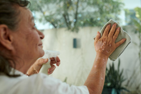 Cropped image of aged woman cleaning glass door with spray detergentの写真素材