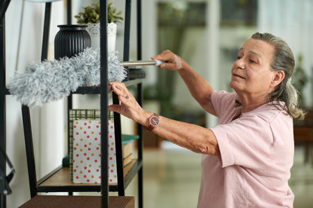 Smiling senior woman wiping off dust from shelves in living room