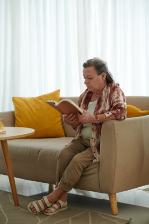 Senior woman sitting on couch in living room and reading bookの写真素材