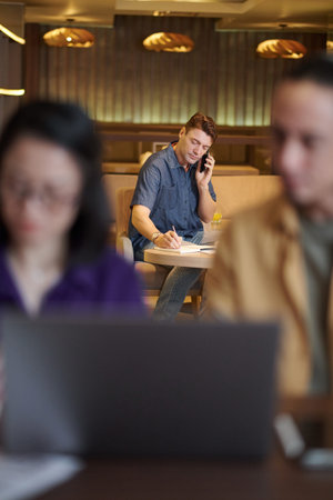 Mature businessman talking on phone with colleague and taking notes in planner when working in...