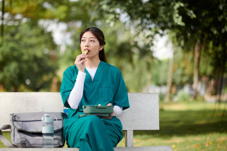Pensive medical nurse in scrubs sitting on bench in park and eating lunchの写真素材