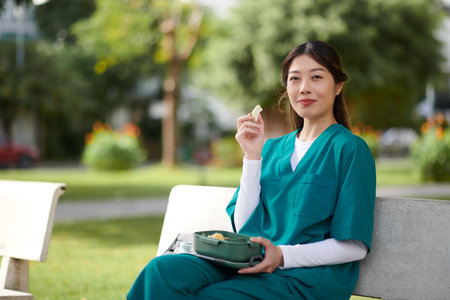 Portrait of cheerful medical nurse eating fresh fruits when resting on bench in park during her breakの写真素材
