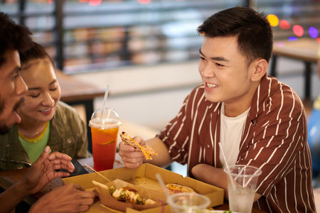 Cheerful Vietnamese young man spending time with friends eating pizza and talkingの写真素材