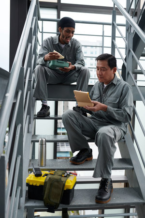 Hungry construction workers sitting on steps and eating sandwiches with coffee for lunch