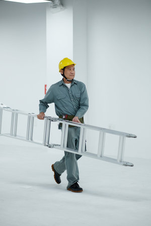 Mature Vietnamese builder in hardhat and overall carrying metal ladder
