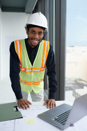 Portrait of smiling construction worker in hardhat leaning on desk with building plan and opened...