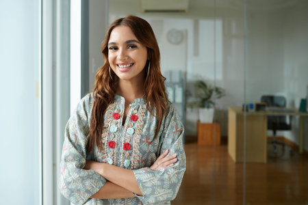 Portrait of young successful female Indian entrepreneur standing by office window